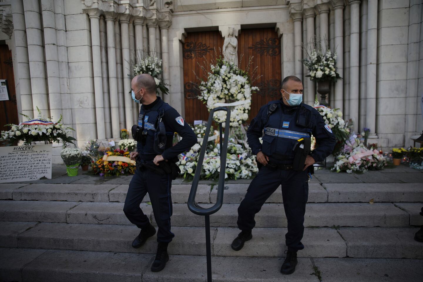 Kommunala poliser står vakt utanför Notre Dame-kyrkan i Nice, Frankrike. Dörrarna och trappan är täckta av vita blommor och kransar.